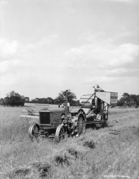 Three-quarter view from front left of a man driving a McCormick-Deering tractor to pull a harvester-thresher in a field.