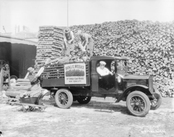 Three men are loading firewood onto the bed of a truck, and one man is sitting in the cab of the truck. There is a large stack of firewood behind the men. The truck has a sign that reads: "Benj. E. Weeks, Firewood,31st Street East River, New York City."