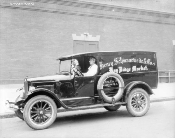 View across street towards a man sitting in the driver's seat of a truck. The sign on the side of the truck reads: "Henry Schwanewede & Co. Inc. Bay Ridge Market. Brooklyn. N.Y." There is a child standing in the passenger seat, and a person is standing behind the truck on a sidewalk in front of a large, brick building.