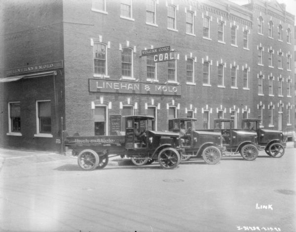 View across street towards men sitting in the driver's seats of four delivery trucks parked at an angle in front of the Linehan & Molo building.