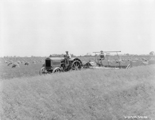 View across field towards two men using a tractor-drawn harvester-thresher.