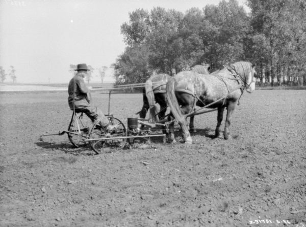 Three-quarter view from right rear of a man using a horse-drawn corn planter in a field.