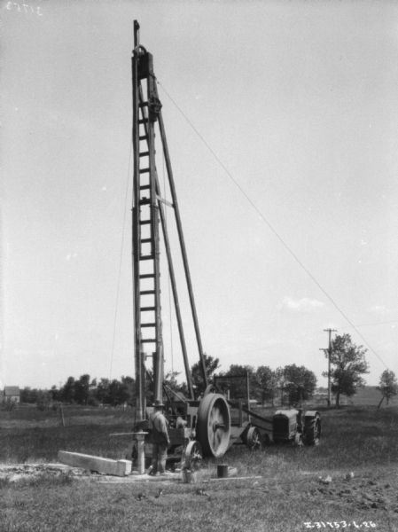View across field towards a man standing near a tractor-powered drill. The tall structure supporting the drill is tethered to the ground.