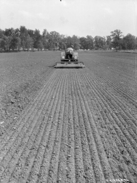 Rear view of a man using a 10-20 tractor to pull a soil pulverizer through a field.