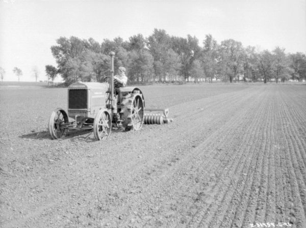 Three-quarter view from front left of a man using a 10-20 tractor to pull a soil pulverizer in a field.