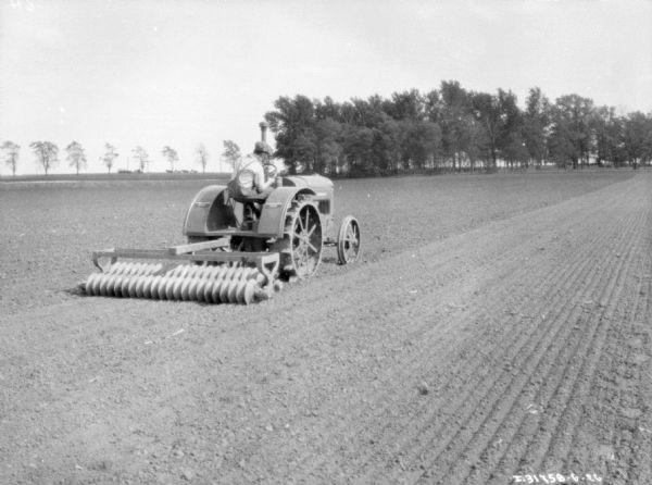 Three-quarter view from right rear of a man using a tractor to pull a soil pulverizer.