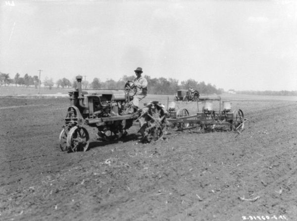 Three-quarter view from front left of a man driving a Farmall tractor to pull a corn planter in a field. In the background is another man driving a tractor.