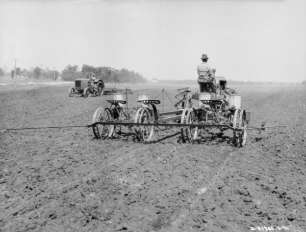 Three-quarter view from right rear of a man driving a Farmall tractor to pull a corn planter in a field. In the background another man is driving a tractor to pull an agricultural implement.