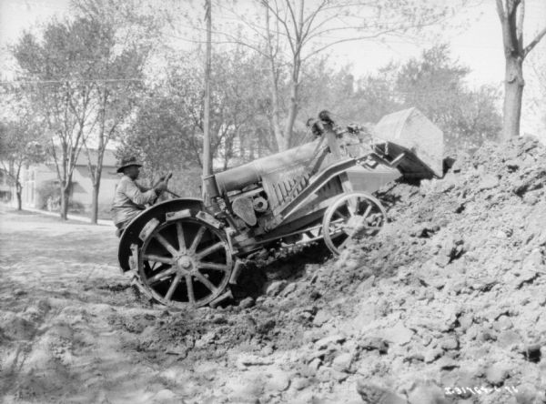 Right side view of a man using a McCormick-Deering tractor to move earth. An unpaved road, and houses among trees are in the background.