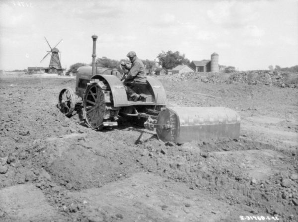 Three-quarter view from left rear of a man using a 10-20 McCormick-Deering tractor to move earth in a field. There is a windmill and farm buildings in the background.