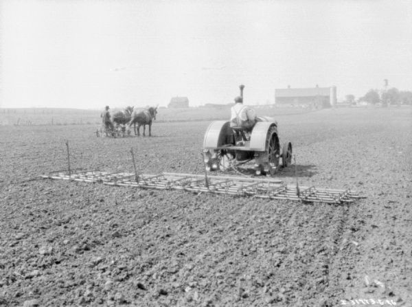 Rear view of a man using a 10-20 tractor to pull a peg-tooth harrow in a field. Another man further down the field on the left is using a team of horses to pull an agricultural implement. Farm buildings are in the distance.