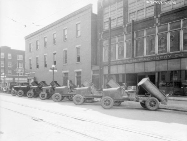 View across street towards a fleet of trucks parked at an angle along the curb. The buildings behind the trucks have signs that read: "Ottawa Farm Machinery Co.," and "College Inn Cafe."