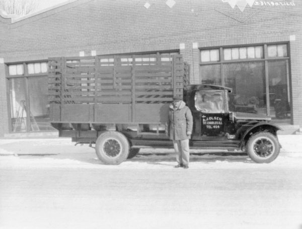 A man is standing in front of the passenger side of a livestock delivery truck parked outdoors. Behind the truck is a brick building with large show windows.