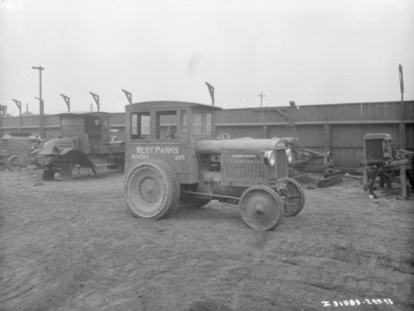 Industrial tractor parked in a yard. In the background are trucks and tractors, and a large, wooden fence.