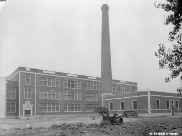 View across yard towards a man using an industrial tractor to move dirt near a factory building with a tall smokestack.