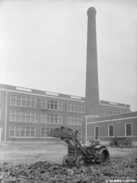 View across yard towards a man using an industrial tractor to move dirt near a factory building with a tall smokestack.