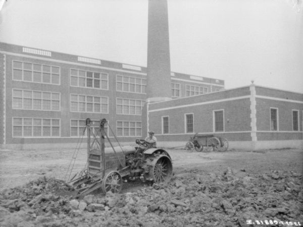 View across yard towards a man using an industrial tractor to move dirt near a factory building with a tall smokestack.