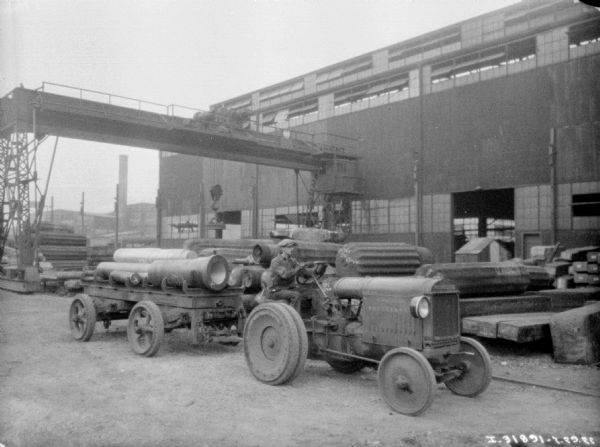 Slightly elevated view of a man using an industrial tractor to pull a wagon carrying parts in a factory yard. 
