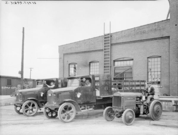 View of a man on a McCormick-Deering industrial tractor parked near two men sitting in the driver's seats of two trucks. The three men are parked  at an angle in front of an industrial building. The signs painted on the sides of the trucks read: "EJ&E Ry. Co." In the background are railroad cars.