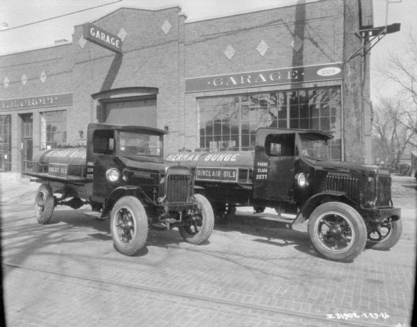 View across cobblestone street towards oil delivery International trucks parked in front of a commercial garage. Signs painted on the side of the trucks read: "Herman Bunge," "Sinclair Oils," and "Phone Elgin 2577." A sign on the garage reads in part: "__L.E. Cropp."