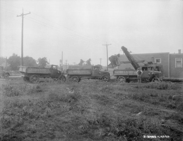 View across field towards men leaning against trucks parked near a steam shovel next to a building.
