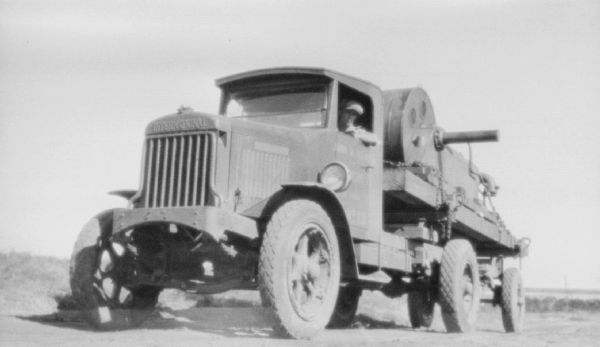 Three-quarter view from front left of a man sitting in the driver's seat of a large industrial truck. There is a large piece of equipment chained to the bed of the truck.