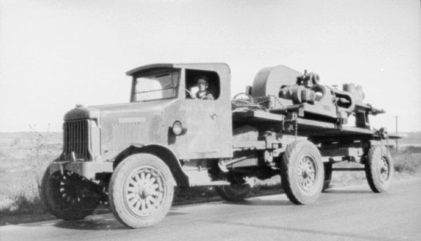 Three-quarter view from front left of a man in the driver's seat of an industrial truck. There is a large piece of equipment chained to the bed of the truck.