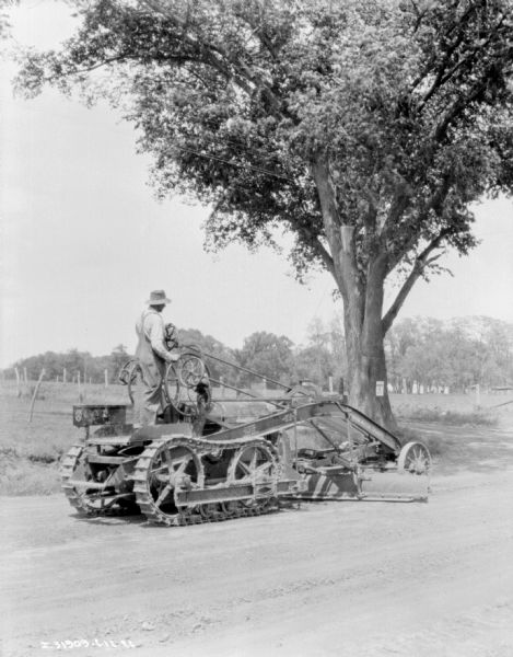 View across unpaved road towards a man operating an Austin Motor Grader.