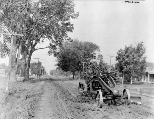 View from front of a man operating an Austin Motor Grader on an unpaved road. There is a house in the background on the right.