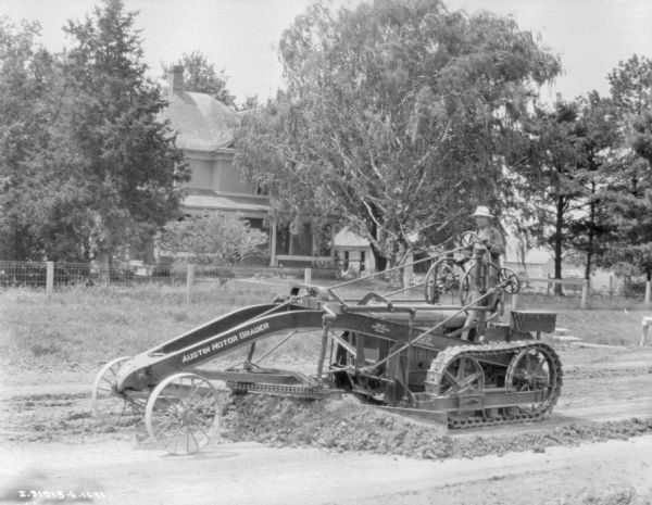 Three-quarter view from front left of a man using an Austin Motor Grader on an unpaved road. There is a house behind a fence in the background.