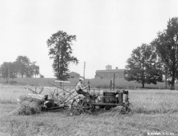 Right side view of a man driving a tractor to pull a binder in a field. There are farm buildings in the distance.