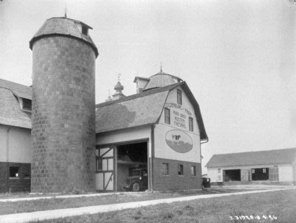 View across barnyard towards a silo next to a barn. A truck is parked inside the open barn door. The sign painted on the side of the barn reads: "Oldenburg • Farm Pure-bred Holstein Friesians."