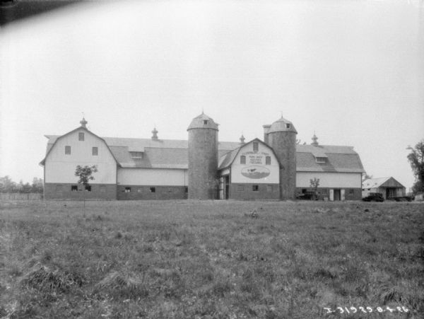 View across field towards silos and large barns. Trucks are parked in the farmyard. The sign painted on the side of the barn reads: "Oldenburg • Farm Pure-bred Holstein Friesians."