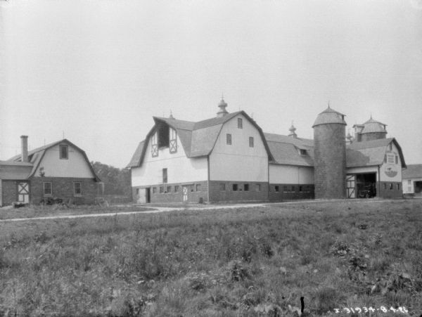 View across barnyard towards a silo next to a barn. A truck is parked inside the open barn door. The sign painted on the side of the barn reads: "Oldenburg • Farm Pure-bred Holstein Friesians."