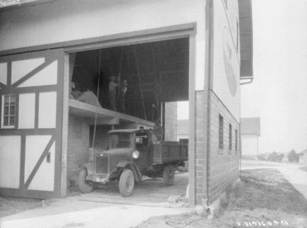 View through open barn door of men loading a truck from a hayloft. Two men are standing in the hayloft, and another man is standing on the bed of the truck.