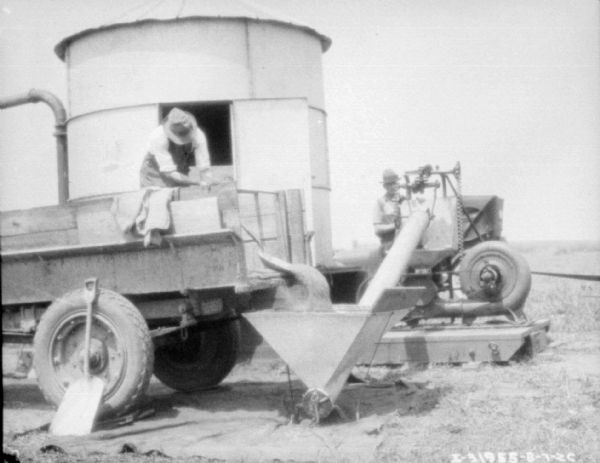 Man standing on truck bed shoveling grain into a hopper. Behind him is a storage building. Another man is standing by machinery on the right.