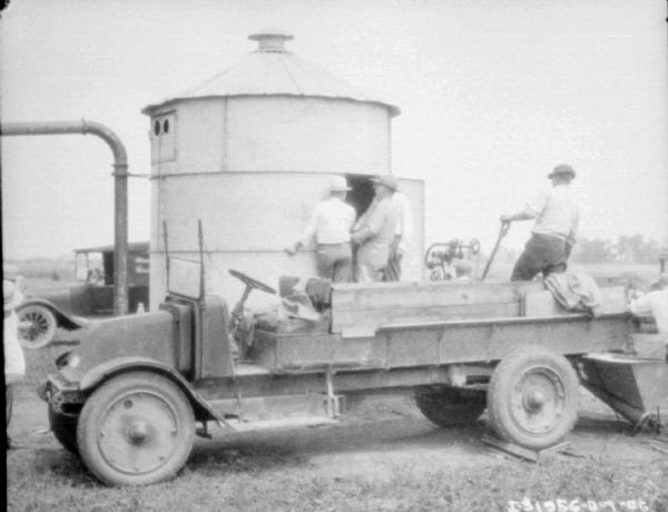 Men are standing on and around a truck in the foreground. Three men are standing at the entrance to a small, round storage building, perhaps for storing grain. There is an automobile and fields in the background.