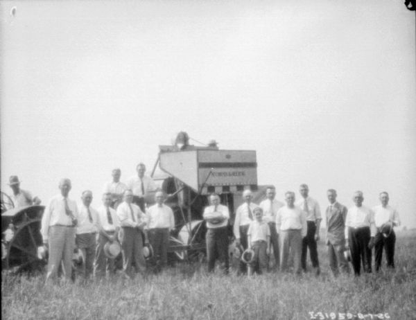 View across field towards a large group of men, and one boy, standing near a tractor, and new demo machinery.