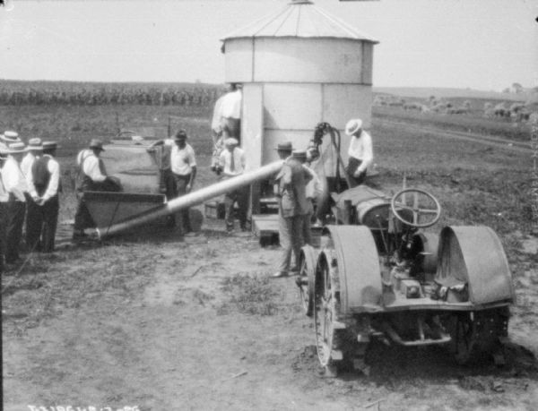 Elevated view of a large group of men gathered around new agricultural demo machinery, a storage building, and a tractor.