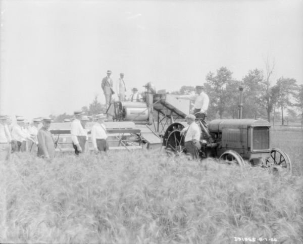 View across field towards a large group of men gathered around new agricultural demo machinery and a tractor.