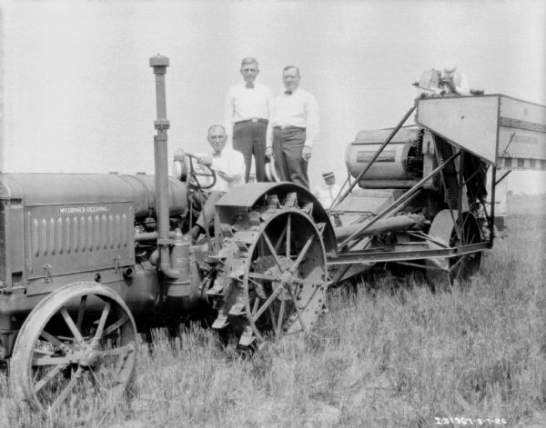Group portrait of a man driving a McCormick-Deering tractor, and two men standing behind him. There is a man standing on the back of the harvester thresher, and other men standing in the field in the background.