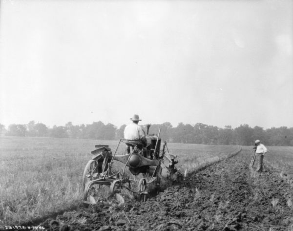 Rear view of a man driving a Farmall to pull a plow in a field. In the background on the right is a man with a movie camera on a tripod.