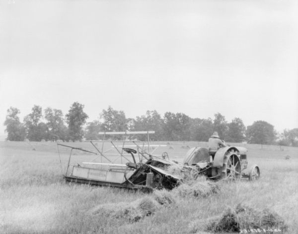Three-quarter view from right rear of a man driving a tractor to pull a binder in a field.