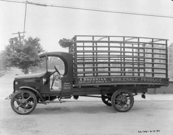 Delivery truck parked outdoors. There are no doors on the cab of the truck. The truck bed has a high stake body, and the sign painted on the truck reads: "A. Brosniak, Wholesale Produce."