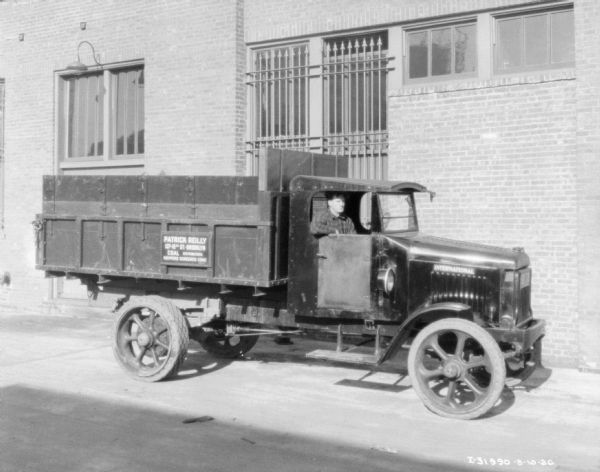 A man is sitting in the passenger seat of a delivery truck parked at the curb in front of a brick building. The sign on the side of the truck bed reads: "Patrick Reilly, Coal Distributors."