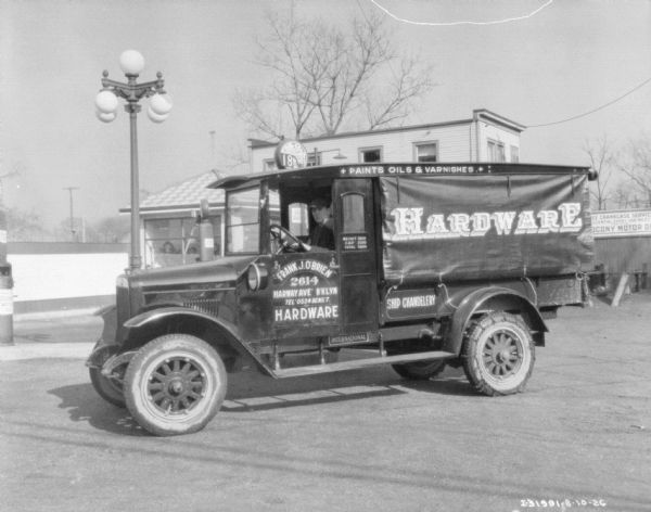 A man is sitting in the driver's seat of a delivery truck parked in front of a service station. The sign on the side of the truck reads: "Hardware, Frank J. O'Brien, Harway Ave, B'klyn."
