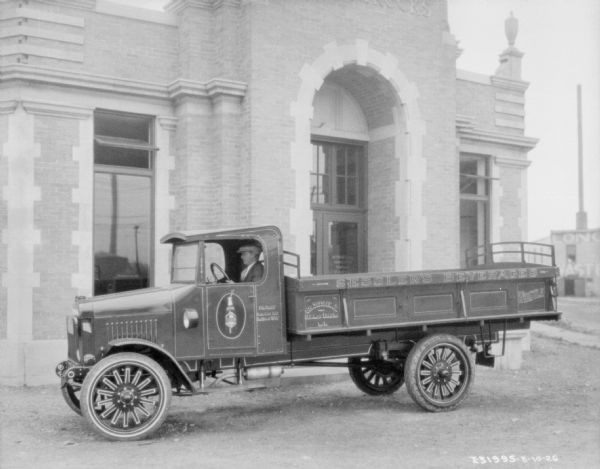 A man is sitting in the driver's seat of an International truck, with signs painted on the side reading: "Sessler's Beverages," "George Sessler, Jr., Glenwood Landing, L.I," and "Whistle." The truck is parked in front of a brick building, with a large archway entrance. A sign on the glass door reads: "International Harvester Company of America."