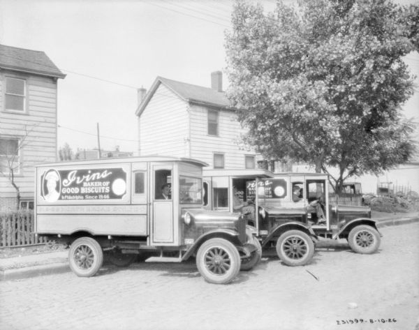 View across street towards three delivery trucks parked in a row at an angle along a curb. A man is sitting in the driver's seat of each truck. Signs on the side of the trucks advertises: "Ivins, Baker of Good Biscuits in Philadelphia Since 1846."