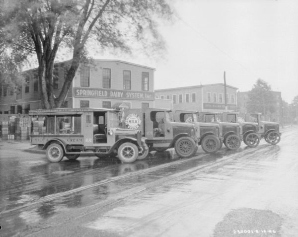 View down rainy street towards a fleet of delivery trucks parked in a row at an angle in front of a building with a sign for: "Springfield Dairy System Inc." Painted on the sides of the trucks are signs that read: "Springfield Dairy System, Milk & Cream."