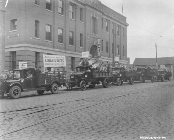 View across cobblestone street towards five trucks on the way to a Kiwanis Race. The trucks are full of men and boys, and have signs that read: "We are on our way to the Kiwanis Races Eastern States Exposition Grounds" and "An After Noon Fun for Everyone." The Boy's Club building is in the background.